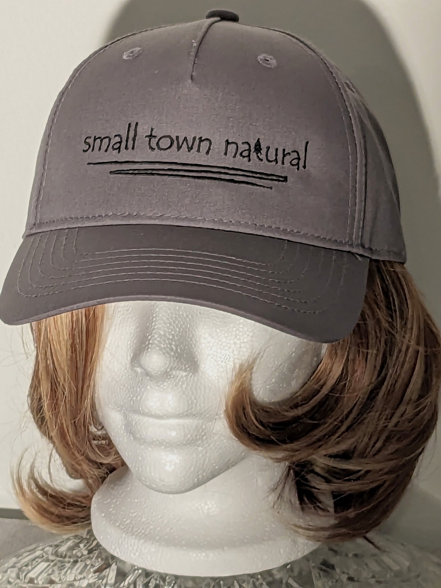 Hat 100% Cotton Small Town Natural ('t' in natural is a tree embroidered graphic of same height as text) is embroidered in plain hand-printed text/font on front of hat in black thread (on black hat thread is light grey) with a thin zigzag line under text representing winding river / creek)