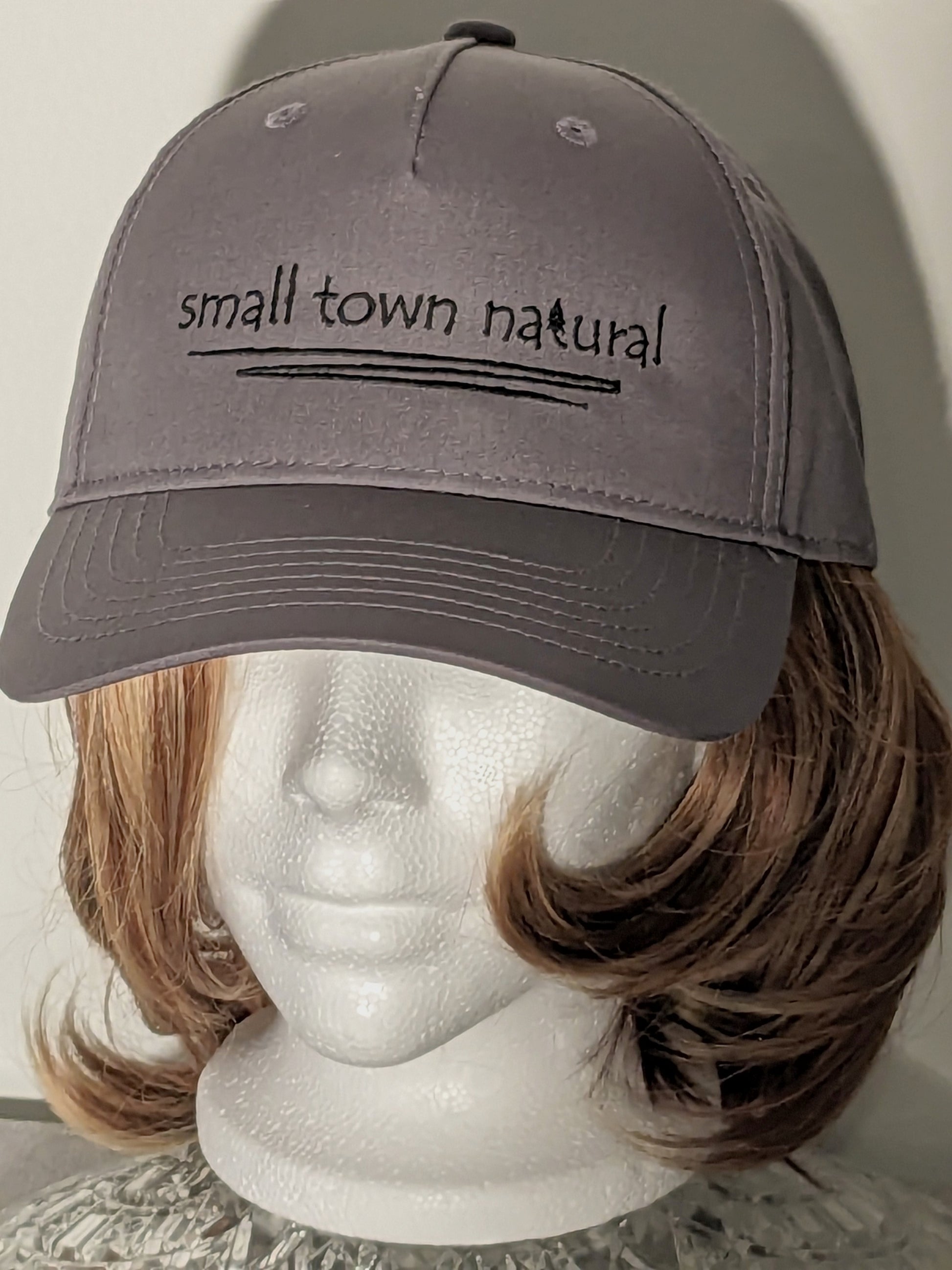 Hat 100% Cotton Small Town Natural  ('t' in natural is a tree embroidered graphic of same height as text) is embroidered in plain hand-printed text/font on front of hat in black thread (on black hat thread is light grey) with a thin zigzag line under text representing winding river / creek)